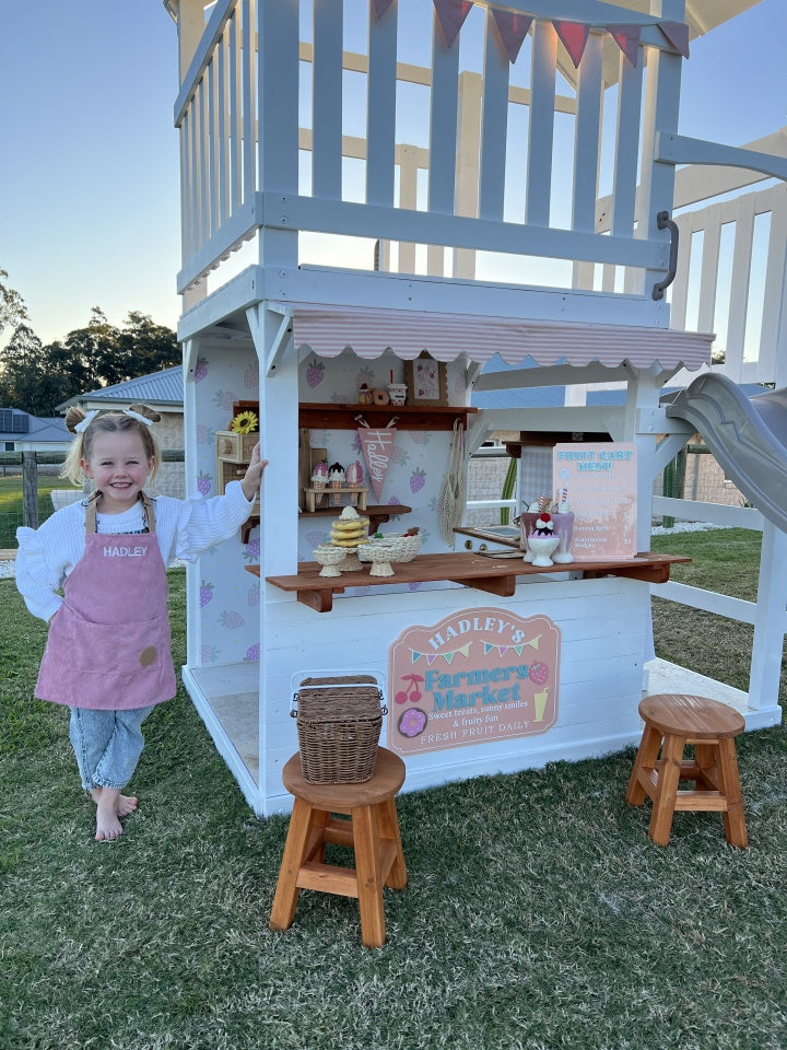 Child in a pink apron standing next to The Rusty Fort by Hide & Seek Kids with a cafe kitchen setup on a grassy area.