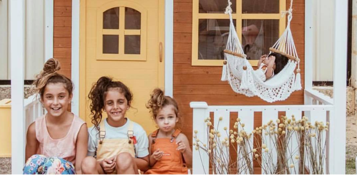 Three children sitting in front of a wooden playhouse with a yellow door.
