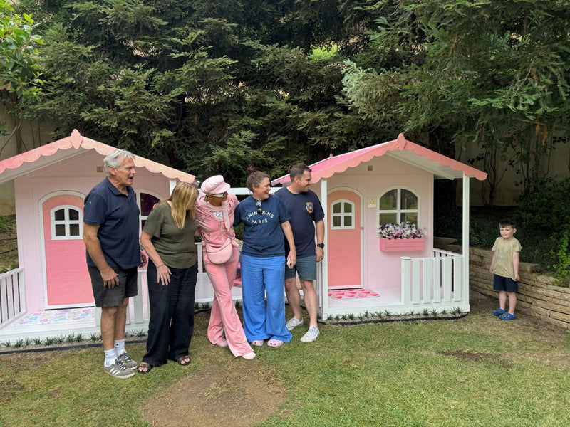 Group of people standing in front of two pink playhouses in a garden setting.
