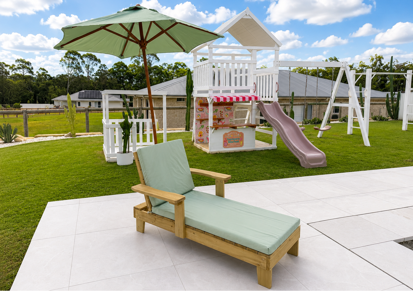 A wooden outdoor chaise lounge with a light green cushion sits on a white tiled patio in front of a backyard play area with a white playhouse, slide, umbrella, and grass under a blue sky.