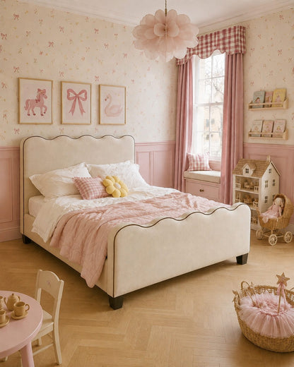 A styled children's bedroom featuring a cream upholstered bed with a scalloped headboard, dressed in pink and white bedding. The room has pink patterned wallpaper, pink wood wainscoting, a pink and white gingham curtain, and various nursery accessories including a dollhouse, basket, and small table.