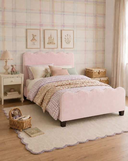 A pink upholstered single bed with a scalloped headboard and footboard, styled in a soft pastel bedroom with plaid wallpaper, cream bedding, a nightstand, and a wicker basket.