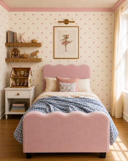 A pink upholstered king single bed with a scalloped headboard and matching footboard, styled in a child's bedroom with pink heart-patterned wallpaper, wooden shelves, a white nightstand, and a blue-and-white checkered blanket.