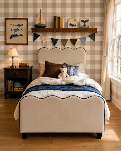 A child's bed with a cream upholstered frame and scalloped edges, styled with white and navy bedding, a teddy bear, and decorative pillows in a room with beige-and-white checkered wallpaper and a wooden shelf.