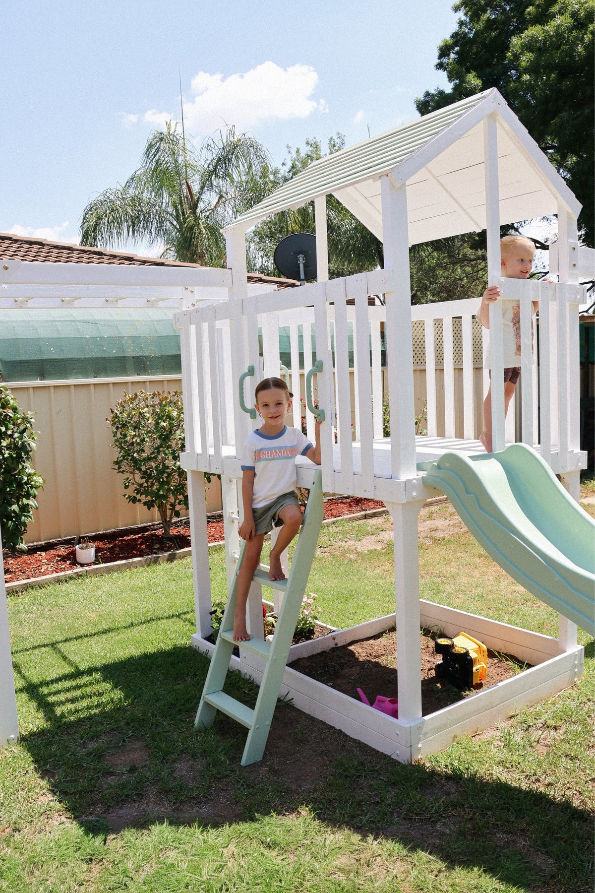 A white wooden house playset with a light green slide and ladder in a backyard, with two children playing on the structure.