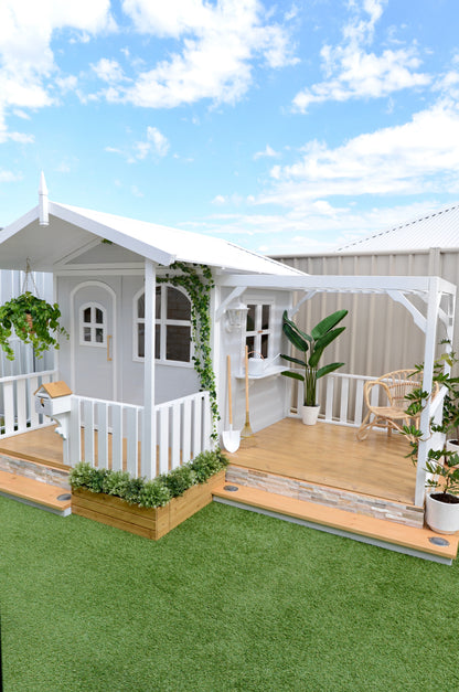 Billie Grand Cubby House by Hide & Seek Kids – grey painted timber cubby with veranda and large arched windows. The Pergola Add-on installed with the cubby house. Flower Box Add-ons surround the cubby house.
