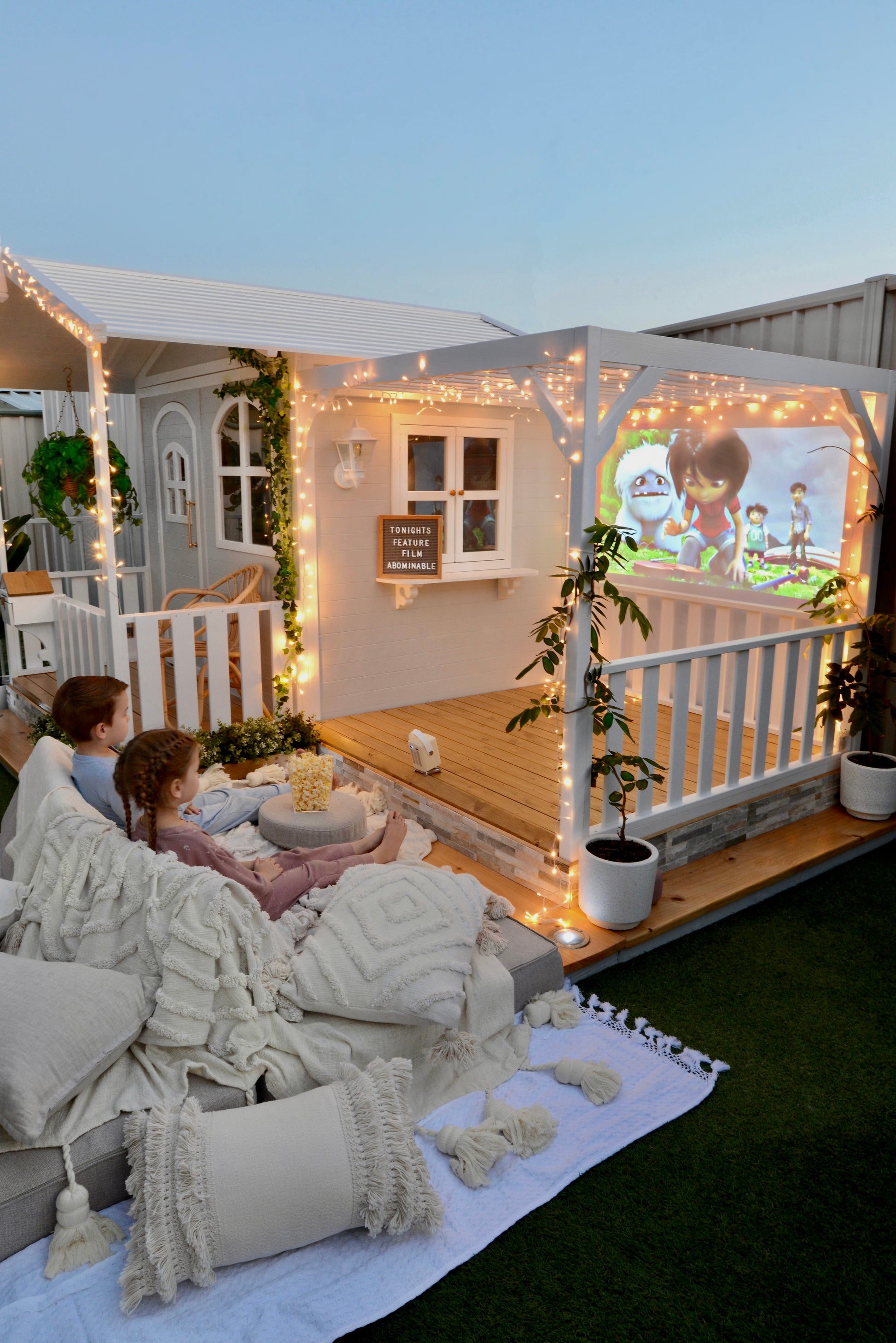 Billie Grand Cubby House by Hide & Seek Kids – Pergola Add-on installed with a cinema setup. Cushions and lights surround the cubby house. Children watching movie.