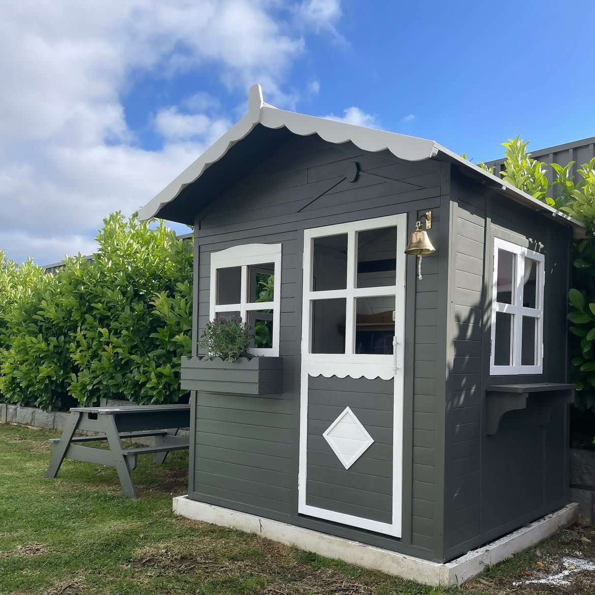 A dark gray wooden garden shed with white trim, featuring a scalloped roof, a front door with a diamond-shaped window, and two side windows. It is situated on a grassy lawn with a wooden bench and green bushes under a blue sky with clouds.