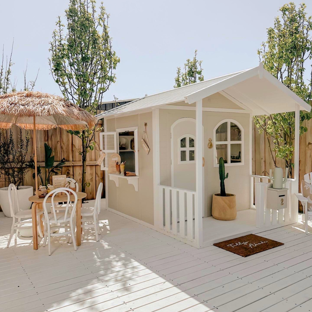 Billie Grand Cubby House by Hide & Seek Kids – Side view of white timber cubby with veranda and large arched windows. Side windows opened, with servery shelf. 