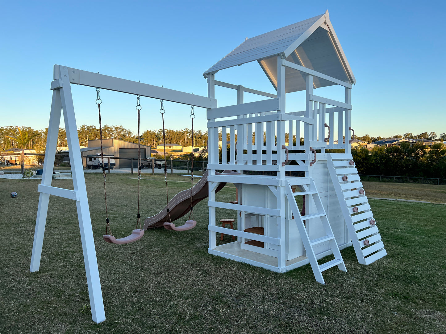 The Rusty Fort Cubby House by Hide & Seek Kids showing climbing wall, rope swings, and elevated playhouse with stairs.