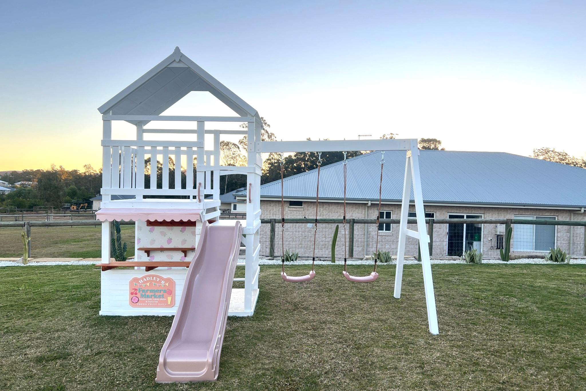 The Rusty Fort Cubby House and Swing Set by Hide & Seek Kids – includes slide, fort, and double swings in backyard setting.