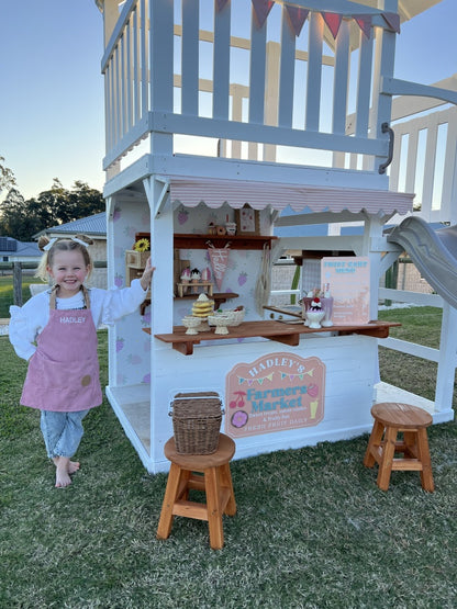 Child in a pink apron standing next to The Rusty Fort  by Hide & Seek Kids with a cafe kitchen setup on a grassy area.